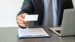 © nunoi - A businessman offers a blank business card while seated at a desk with a laptop and documents nearby.