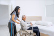 © Creativa Images - Female Caregiver Pushing Senior Man in Wheelchair Next to Bed in Bright Room