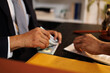 © Kield KD - A man is handing a stack of money to another man. The man receiving the money is wearing a suit and tie. The scene appears to be a business transaction or negotiation