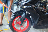A person using a high-pressure nozzle to spray water on a scooter's black tire and vibrant red alloy rim, cleaning the front wheel during a wash.