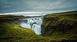 Dramatic waterfall cascades over rock ledges, lush green fields under a moody sky