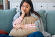 © Srdjan - Young woman at home hugging pillow on the sofa feeling unwell with abdominal discomfort possible menstrual cramps stomach ache food poisoning or digestive upset resting under blanket, seeking relief