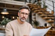 © Gwendolyn Davis - A focused and distinguished mature man, wearing stylish eyeglasses and a comfortable knit sweater, gazes thoughtfully directly at the camera while holding a paper document. His salt-and-pepper hair an
