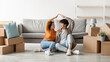 © Prostock-studio - A happy Asian couple enjoys their moving day as they sit among cardboard boxes in their new home. They are making a house roof gesture, symbolizing their fresh start together.