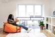 © Prostock-studio - A woman enjoys a quiet moment reading an open book while sitting comfortably in a vibrant chair. Sunlight streams through large windows, illuminating the room.