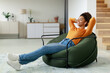 © Prostock-studio - A young woman relaxes in a modern chair at home, wearing earbuds and smiling. She is dressed casually in an orange sweater and jeans, enjoying a moment of peace.