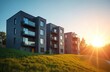 © Maryna - Modern multi-story apartment buildings with dark brick facades stand on green grassy slope. New residential complex features sleek glass balconies, reflecting golden sun. Bright sun shines creating