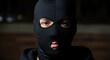 © Chandara - A person wearing a black balaclava, with a neutral expression, standing in a dimly lit room with a wooden floor and a dark background.