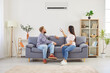 © Studio Romantic - Young family couple sitting on sofa in living room and turning on air conditioner. Happy man and woman using remote control and setting up comfortable temperature on AC system to cool down their home.