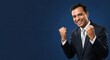 © Nina - Happy Indian businessman in a dark suit celebrating success with a triumphant gesture against a blue background.
