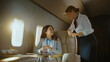 © Videophilia - Businesswoman relaxing in a private jet cabin, holding a champagne glass, and smiling while interacting with a friendly flight attendant providing premium service during her luxurious air travel