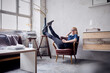 © TrueShot | Westend61 - Happy woman sitting with legs in the air on armchair in loft