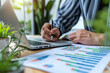 ©  𝙿𝚒𝚡𝚎𝚕𝙶𝚕𝚘𝚠  - Close up of a person s hands writing on a document with a pen while working on a laptop computer with charts and graphs on the desk illuminated by natural light