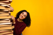 © deagreez - Young mixed race woman smiles while stacking a tall pile of book against a bright yellow background for study and discovery