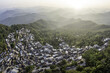 © AmazingAerialAgency - Aerial view of a village nestled in lush green hills, with houses tightly packed together, contrasting with the distant hazy mountain ranges, Menghong Village, Yunnan, China.