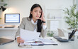 © wattana - Young businesswoman rubbing her temple while reading financial documents at a messy desk—calculator, charts, and paperwork—office stress concept.