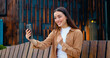 © serg - Close up of pretty Caucasian girl talking on video call using phone outdoors while sitting on bench near office building. Online chat
