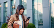 © serg - Young Caucasian woman confidently walking near a modern glass office building, smiling as she carries a laptop and a notebook in her hands, while checking her smartphone.
