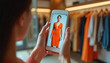 © miss irine - Woman uses smartphone in fashion store. She views orange dress on phone screen. Modern boutique showcases virtual fitting room tech for shoppers. Technology and fashion combine in retail scene.