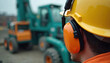 © Viktor - Man wears yellow hard hat, orange ear defenders on noisy construction site. Heavy machinery like diggers, loaders operates near worker. Safety gear protects from loud sounds, impacts during work.