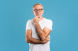 © Prostock-studio - Mature man standing alone against a blue background is deep in thought. He wears glasses and a white t-shirt while touching his chin, contemplating an important decision.
