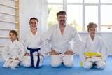 Smiling parents with preteen children, son and daughter, wearing traditional white kimonos sitting on knees on tatami in gym, ready for martial arts training. Sports family concept
