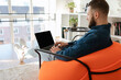 © Prostock-studio - A young man sits comfortably in an orange chair while working on his laptop. Sunlight fills the modern room, highlighting the cozy atmosphere and minimalistic decor.