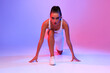 © Prostock-studio - Fit woman in a crouch start position looks at the camera, preparing for her running workout. She is set against a vibrant pink and blue neon studio background, showcasing her determination.