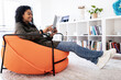 © Prostock-studio - A woman sits comfortably in an orange chair, engaging with her tablet. She enjoys the cozy atmosphere of a bright living room filled with books and decor during the day.