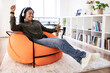 © Prostock-studio - A young woman smiles and dances slightly while wearing headphones in a cozy and bright living room. She is seated in an orange bean bag chair surrounded by bookshelves and plants.