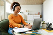 © Prostock-studio - A young woman wearing headphones smiles as she works on her laptop at a stylish table. She is surrounded by a cozy atmosphere, with plants and stylish decor.