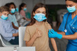 © Prostock-studio - Girl wearing a face mask smiles and gives a thumbs up as a nurse places a bandage on her arm. Other children wait in the background at a vaccination clinic during coronavirus immunization.