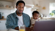 © Helga - Young African-American father working remotely from home while holding his baby and sitting with his son in a bright modern kitchen. The father looks focused on his laptop, with a
