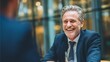 © Eve - Close-up of a charismatic senior businessman in a suit laughing genuinely during a successful one-on-one business meeting or corporate interview in a modern setting.
