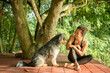 © sutlafk - A happy Asian woman in yoga attire sitting cross-legged on a wooden deck, smiling warmly at her fluffy, gray dog. They are under a large tree with lush green foliage in the background.