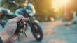 © ds17 - A hand holding motorcycle keys in the foreground with a motorcycle parked in the background during sunset. The scene captures a moment of anticipation and freedom.