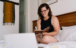 © BullRun - Confident middle-aged woman writing notes while working remotely from bed, multitasking between digital screen and paper to stay organized and productive in casual home setting.