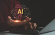 © MUNTHITA - Close up of hand holding glowing AI hologram above laptop, symbolizing artificial intelligence, innovation, digital transformation, technology development and futuristic business concept in modern era