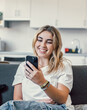 © Daniel - Happy pleasant millennial girl relaxing on comfortable couch, holding smartphone in hands. Smiling young lady chatting in social networks, watching funny videos, using mobile applications at home.
