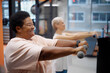 © Seventyfour - Caucasian senior woman and Black middle aged woman exercising with dumbbells in gym, both extending arms forward while focusing on strength training during fitness session