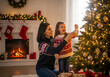 © shirish - Happy mother and daughter decorating a Christmas tree with ornaments in a festive living room. Cozy fireplace with stockings and holiday lights in the background. Family tradition.