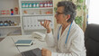 © Krakenimages.com - Senior woman doctor with grey hair in a clinic office, wearing a stethoscope, using a smartphone to send a voice message while working on a laptop.