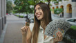 © Krakenimages.com - Woman holds bitcoin coin and a fanned stack of dollars while smiling on city street; financial success.