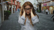© Krakenimages.com - Young caucasian woman with red hair and glasses holds face with both hands on narrow city street amid blurred buildings; stress.