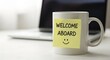 © Shabbir - Bright welcome aboard note with smiley face on coffee mug beside laptop in modern office setting, creating a warm and inviting atmosphere for new team members.