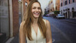 © Krakenimages.com - Young woman grinning widely and clasping hands in front of brick buildings on a bustling city street; happiness.