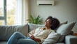 © Pete - Young black woman smiles relaxing on couch. Home air conditioner provides cool comfortable indoor climate. She enjoys fresh air flow in living room during hot summer day.