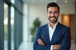© Pete - Attractive Latin business man in navy suit. Confident entrepreneur smiles at camera. Professional model stands in modern office with crossed arms.