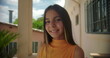 © Marco - Close-up portrait of a smiling young girl on a Latin American balcony with pastel buildings and greenery in the background, cheerful and confident expression