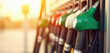 © Pete - Colorful gas pump nozzles line up at fuel station. Cars refuel with gasoline or diesel. Workers help drivers pay for service. Energy prices rise.